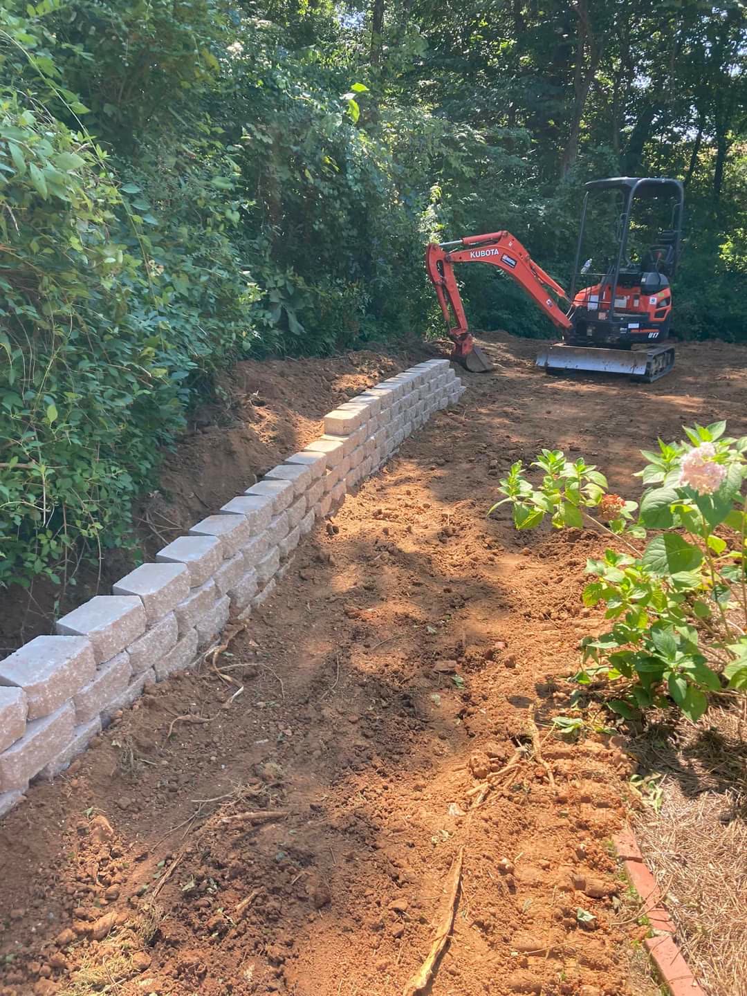 Block retaining wall being built with an excavator along a tree line, Chattanooga TN area